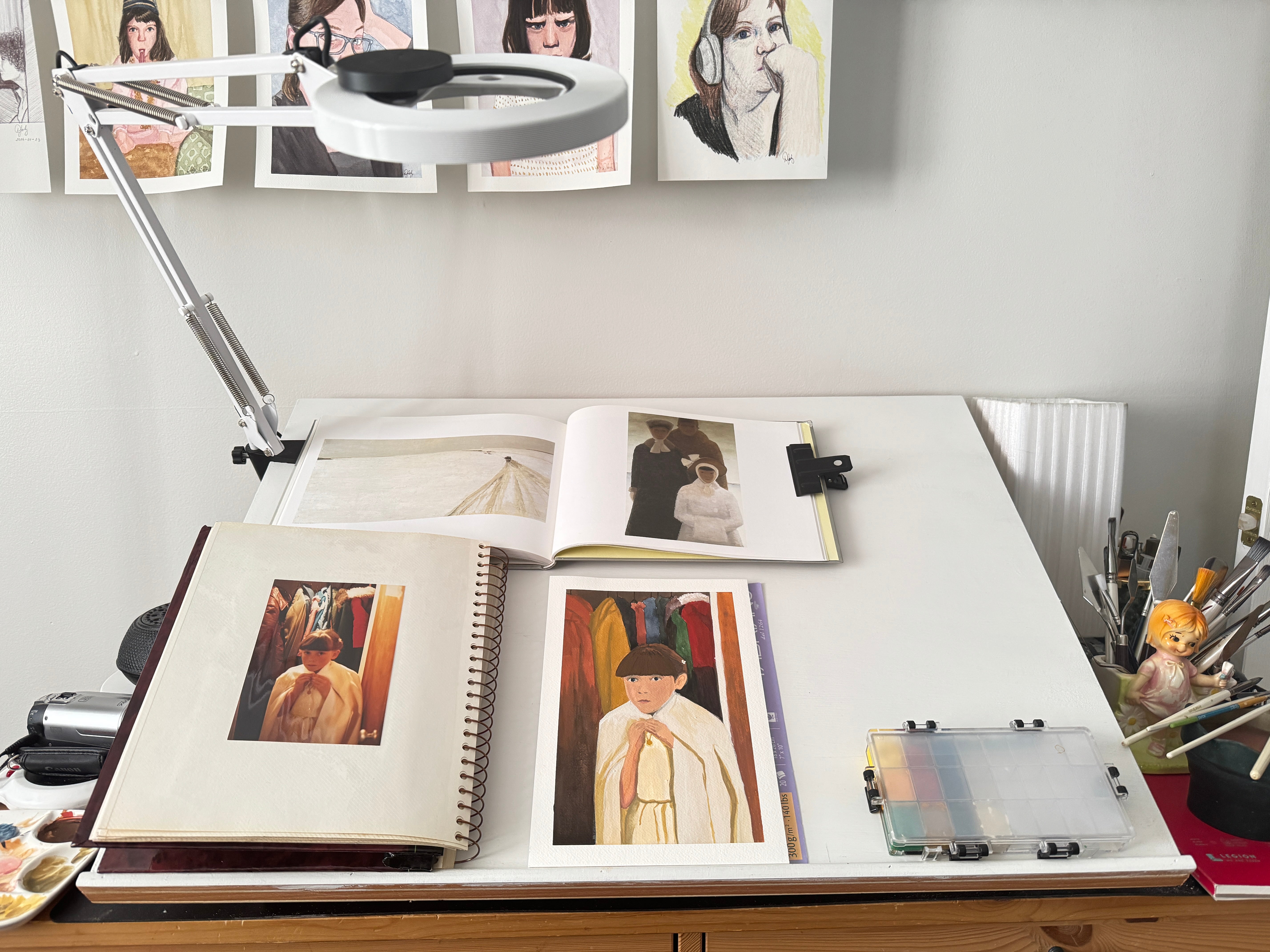 Open photo album on a desk with a lamp above, showing images of people and a portrait painting in process.